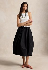 Γυναίκα που φοράει λευκό τοπ χωρίς μανίκια, μαύρη φαρδιά μεσαίου μήκους φούστα, διακριτικά στρωμένα μπλε και κίτρινα κολιέ, και κόκκινες σαγιονάρες, στέκεται με σταυρωμένα χέρια.