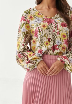 Woman wearing long-sleeve floral blouse with puffed sleeves and pink pleated skirt, hands gently clasped at waist.
