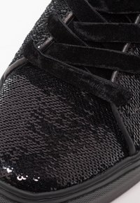 Bout de sneaker noire avec des sequins brillants et trois lanières en velours doux traversant les lacets sur un fond blanc.