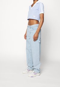 Polo cropped azzurro chiaro con colletto bianco e dettagli ricamati, abbinato a jeans azzurro chiaro a vestibilità ampia e scarpe da ginnastica bianche con accenti viola.