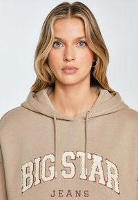 Béžová mikina s klokaní kapsou vpředu, na které je vyšívaný nápis "BIG STAR" v texturovaných krémových a vínových písmenech. Nastavitelná kapuce se stahovací šňůrkou.