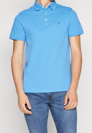 Person trägt ein hellblaues, kurzärmeliges Poloshirt mit Knöpfen und einem kleinen Logo auf der Brust, kombiniert mit blauen Jeans.