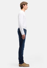 Granatowe, obcisłe jeansy o kroju slim z pięcioma kieszeniami, szczegółowym przeszyciem, noszone w połączeniu z białą koszulą z długim rękawem i beżowymi botkami sięgającymi kostek. Widok z boku.