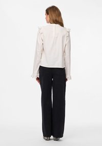 Blusa blanca con acentos de volantes en los hombros y detalles de encaje en las mangas, combinada con pantalones anchos negros y zapatos con estampado.