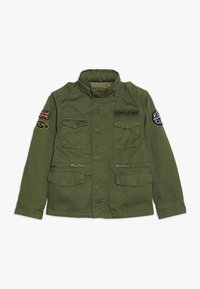 Oliwkowa kurtka z bawełny, z kołnierzykiem stójkowym, dwoma przednimi kieszeniami oraz różnymi naszywkami, w tym brytyjską flagą i logo.