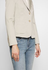 Veste beige cintrée avec deux poches avant et boutons, portée avec un jean en denim bleu clair.