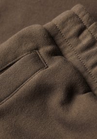 Tela de algodón marrón con una textura suave, con costuras visibles y un detalle de bolsillo ribeteado. El material parece ligero y transpirable.
