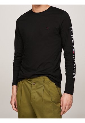 Homme portant une chemise noire à manches longues avec un petit logo Tommy Hilfiger sur la poitrine et le texte « Tommy Hilfiger » sur la manche droite, associée à un pantalon vert olive.