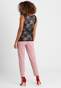 Černý krajkový top s květinovým vzorem, spojený se světle růžovými kalhotami na míru a červenými vysokými podpatky. Pohled zezadu s texturovaným účesem.