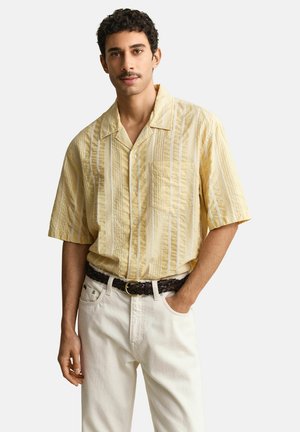 Mann trägt ein gelb gestreiftes kurzärmeliges Hemd, das in weiße Hosen mit einem geflochtenen braunen Gürtel gesteckt ist, und steht mit einer Hand in der Tasche.