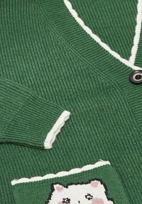 Grön stickad kofta med vit kant, med en svart knapp och en fickdesign av en seriekatt med rosa detaljer.