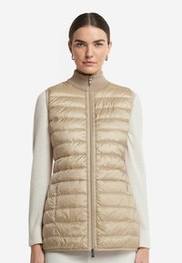 Beige pufferväst med vertikal quiltad textur, hög krage och framfickor. Bärs över en ljusfärgad tröja.