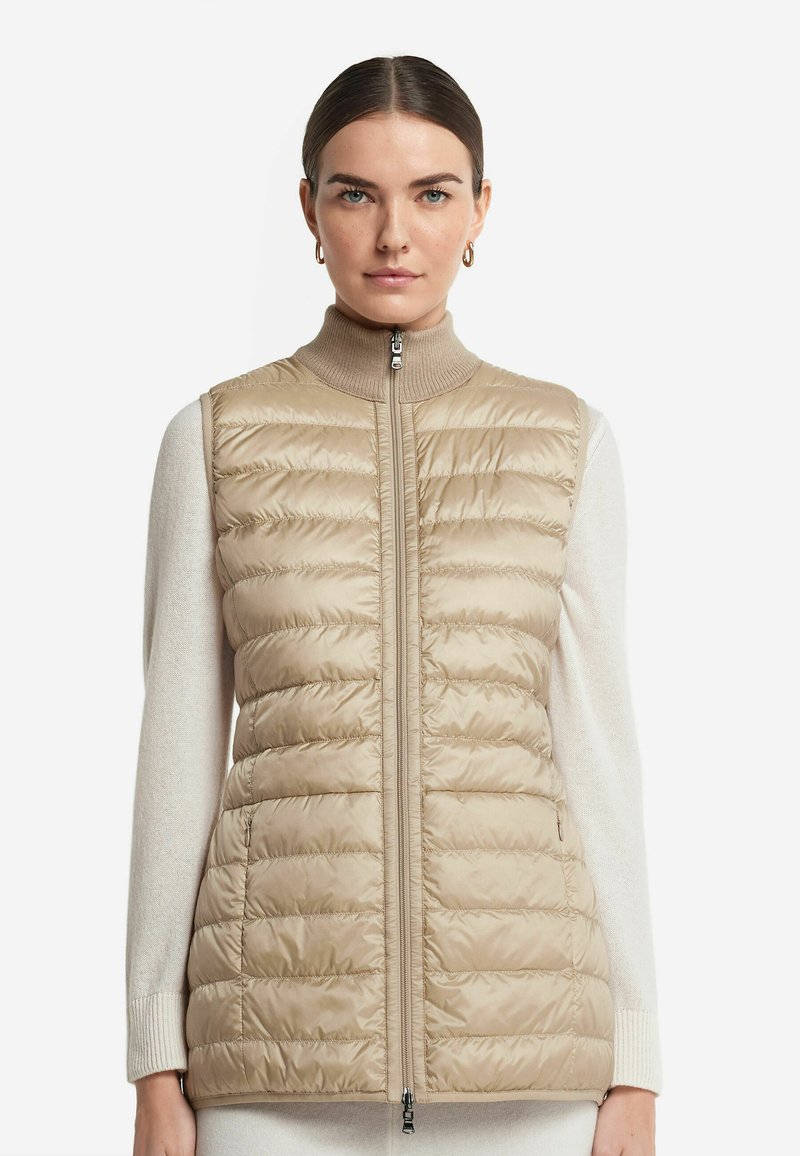 Beige pufferväst med vertikal quiltad textur, hög krage och framfickor. Bärs över en ljusfärgad tröja.