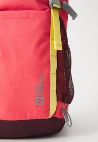 Pink fabric backpack with a yellow strap and gray accents. Features mesh pocket and silver logo. Textured surface with dark base.