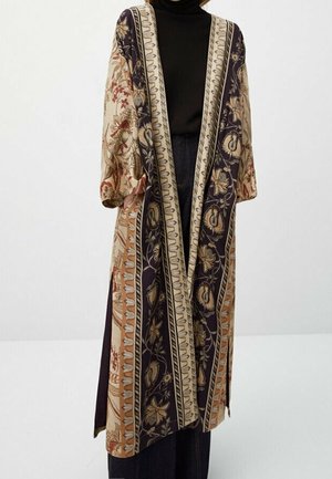 Długi kardigan w stylu kimono z otwartym przodem, zdobiony kwiatowymi i geometrycznymi wzorami w odcieniach beżu, czerni i brązu, noszony na czarnym golfie i ciemnych dżinsach.