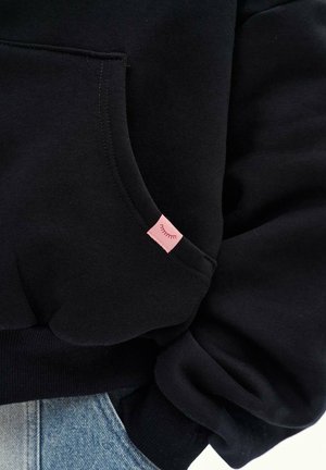 Czarna bluza z kapturem z kieszenią i małą różową metką z ikoną zamkniętej powieki, noszona na jasnoniebieskich dżinsach.