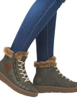 Jambes en jean bleu portant des bottes d'hiver hautes grises avec des lacets beiges, des semelles en caoutchouc marron, une fermeture éclair latérale et une bordure en fourrure autour de la cheville.