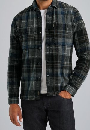 Man wearing dark plaid button-up shirt over white T-shirt with dark jeans, one hand in pocket, against plain gray background.