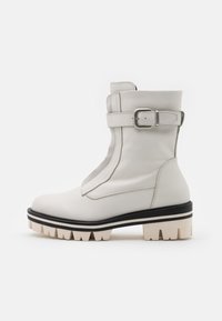 Botas blancas hasta el tobillo con un acabado de cuero texturizado, que presentan una correa y hebilla prominentes. Suelas de goma gruesa con un diseño contrastante.