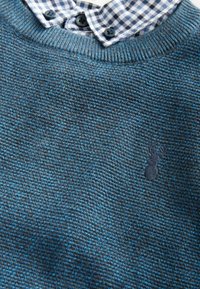 Sweter robiony na drutach w ciemnoniebieskim i szarym kolorze o teksturowym wykończeniu. Posiada kołnierzyk w kratkę z dwoma guzikami oraz mały wyszywany logo ananasa.