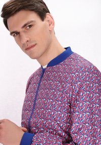 Giacca bomber a fantasia con colletto e polsini blu, caratterizzata da un design vivace di forme intrecciate rosa e bianche su uno sfondo blu.
