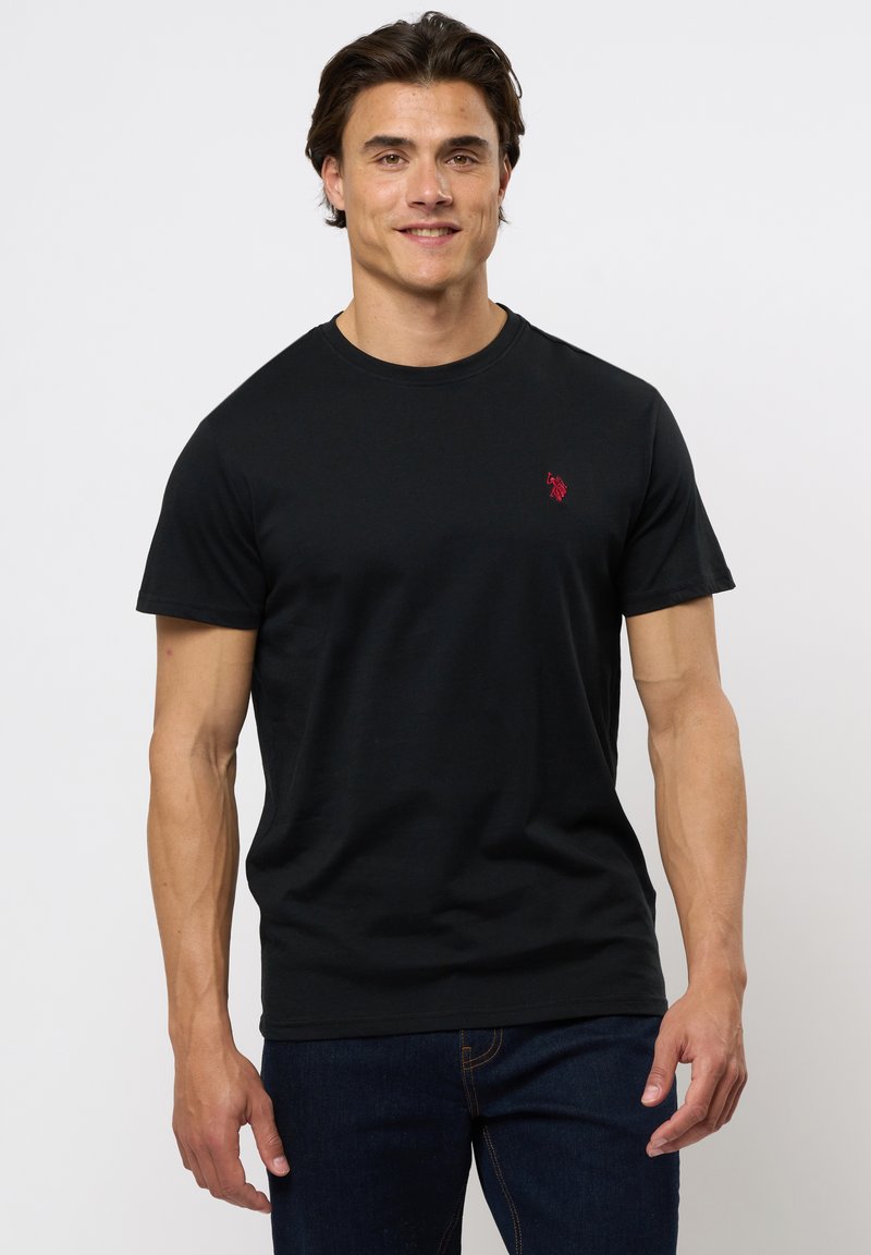 Tricou din bumbac negru cu guler rotund și mâneci scurte. Prezintă un mic logo roșu pe piept. Este purtat cu blugi de denim de culoare bleumarin.