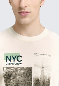 Trumpas rankovės kreminis marškinėliai su NYC tekstu tamsiai mėlynos ir žalios spalvų, vaizduojantis pastato grafiką ir pikselinį dizainą. Pagamintas iš medvilnės, su apvaliu kaklu.