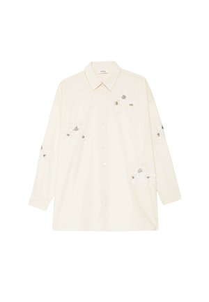 Chemise crème à manches longues avec col et boutons, ornée de broderies florales blanches aux centres noirs sur le devant, les manches et les côtés inférieurs.