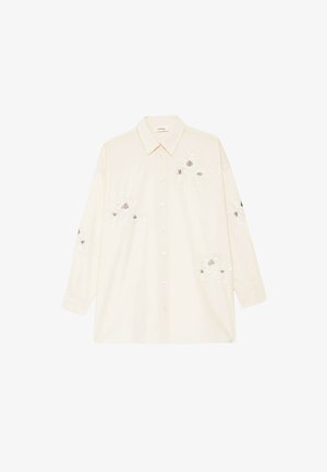 Chemise crème à manches longues avec col et boutons, ornée de broderies florales blanches aux centres noirs sur le devant, les manches et les côtés inférieurs.