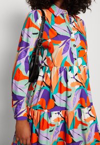 Robe colorée et à motifs présentant des feuilles orange, violettes, vertes et blanches, avec un col et une jupe à volants. Un accessoire à bandoulière noire est visible.