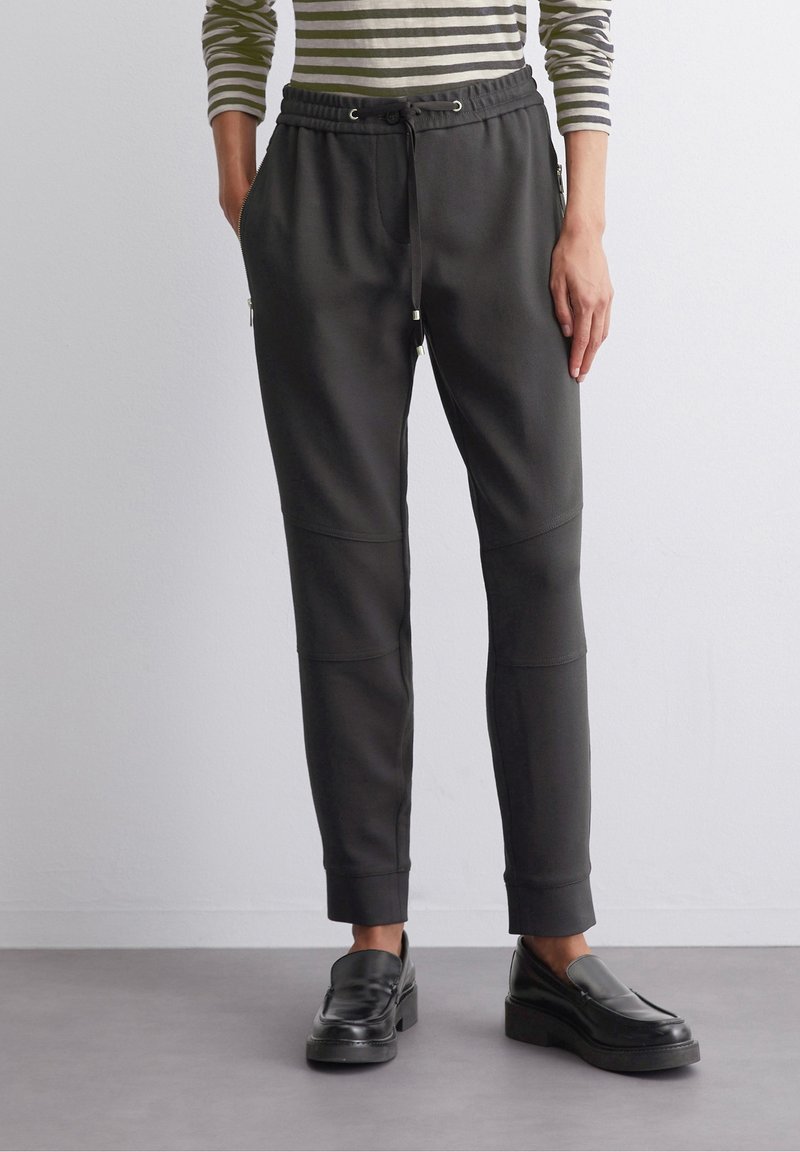 Pantalones jogger negros con cintura ajustable con cordón, puños acanalados y bolsillos laterales con cremallera. Textura suave con detalles en paneles. Combinados con zapatos negros sin cordones.