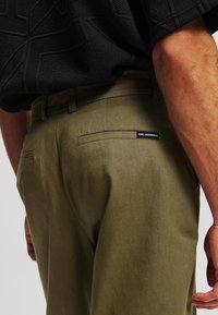 Pantaloni verde oliva con una texture liscia, dotati di una tasca posteriore con etichetta del marchio e un design a gamba dritta.