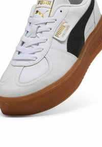 Białe skórzane sneakersy z czarnym paskiem po boku, płaskimi sznurówkami i tanową gumową podeszwą. Posiadają złote logo na języku i boku.