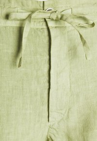 Pantalon en lin vert clair avec une ceinture en tissu nouée en nœud à la taille, avec une texture et des détails de couture visibles.