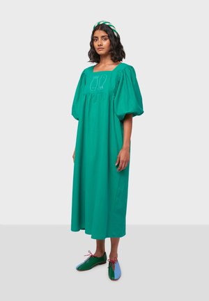 Abito in cotone verde con maniche a sbuffo e scollatura quadrata, caratterizzato da dettagli ricamati. Abbinato a scarpe bicolore verdi e azzurro chiaro.