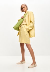 Completo blazer e pantaloni corti in lino giallo chiaro, abbinato a una maglietta beige e scarpe slip-on intrecciate. Tieni in mano una borsa a tracolla verde.