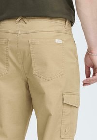 Khaki spodnie cargo wykonane z bawełny, z wieloma kieszeniami, prostymi nogawkami oraz metką na tylnej kieszeni. Gładka tekstura.