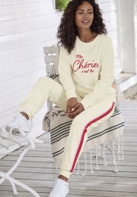 Cremefarbene Loungewear-Set mit rotem Text und gestreiften Akzenten, bestehend aus einem Sweatshirt mit Rundhalsausschnitt und schmal zulaufenden Hosen, kombiniert mit weißen Sneakers.