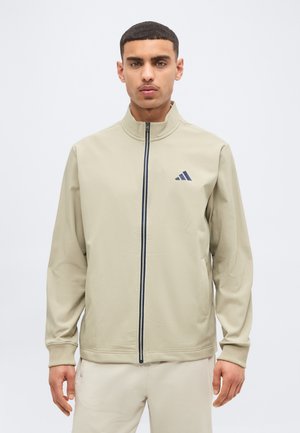 Άνδρας που φοράει μπεζ ζακέτα με πλήρες φερμουάρ και όρθιο γιακά, με σκοτεινό λογότυπο στο αριστερό μέρος του στήθους, συνδυασμένη με μπεζ παντελόνι, κοιτάζοντας μπροστά.