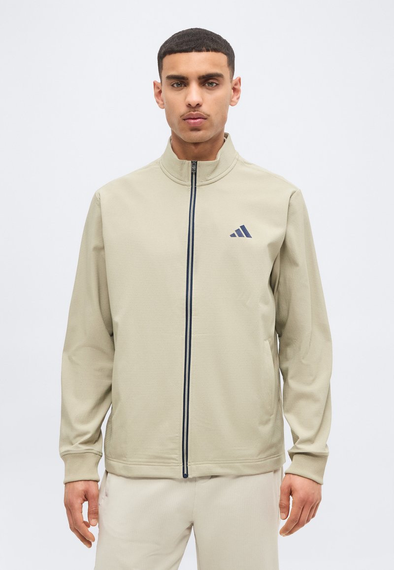 Άνδρας που φοράει μπεζ ζακέτα με πλήρες φερμουάρ και όρθιο γιακά, με σκοτεινό λογότυπο στο αριστερό μέρος του στήθους, συνδυασμένη με μπεζ παντελόνι, κοιτάζοντας μπροστά.