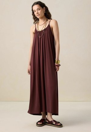 Maroon maxi dress with spaghetti straps, gathered detail at the neckline, and a flowing silhouette. Paired with strappy sandals and accessories.
