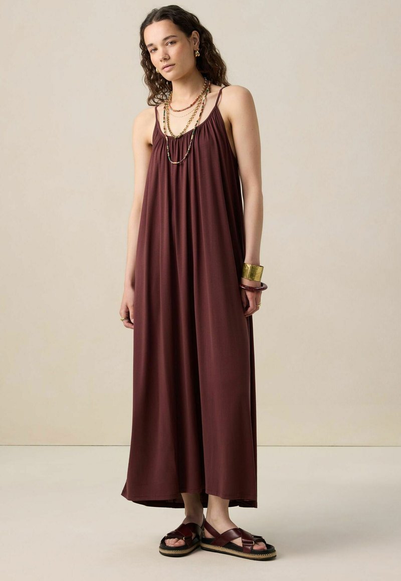 Robe maxi bordeaux avec des bretelles spaghetti, un détail froncé au niveau du décolleté et une silhouette fluide. Associée à des sandales à lanières et des accessoires.