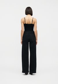 Γυναίκα που στέκεται με την πλάτη, φοράει μαύρο αμάνικο τοπ και μαύρο ψηλόμεσο παντελόνι με φαρδιά ποδόγυρα και διακριτικό μοτίβο σε λευκό φόντο.