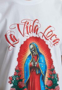 Balts T-krekls ar krāsainu grafiku, kurā attēlots skeleta figūra z blue apmetni, sarkanu kleitu, ar rozēm un tekstu "La Vida Loca".