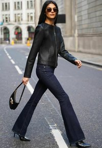 Black leather jacket, dark blue flared jeans, black stiletto heels, and a small black handbag, shot on a city street with asphalt surface.
