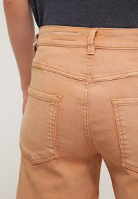 Pantalons en denim orange clair avec taille haute, deux poches arrière et détails de coutures visibles. Le tissu a une texture lisse.