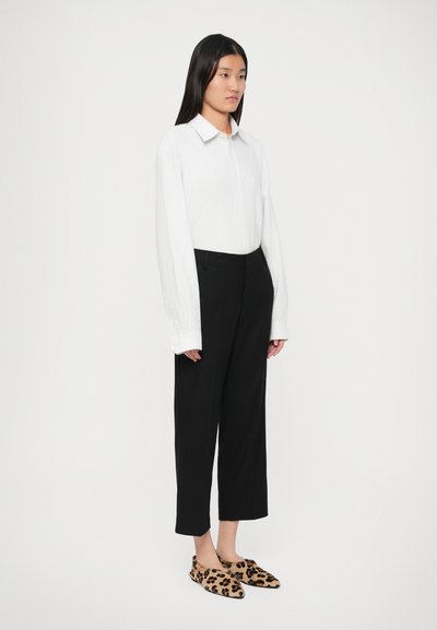 Chemise blanche à manches longues avec col, pantalon noir taille haute et chaussures à enfiler à imprimé léopard. La tenue présente une coupe décontractée et une texture lisse.
