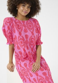 Roza obleka z rdečimi vezenimi vzorci, napihnjenimi rokavi in sproščenim krojem. Gladka tekstura, do kolen, z diamanti in cvetličnimi detajli.