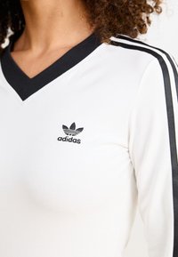 Biała koszula z dekoltem w serek z czarnymi akcentami, z czarnym logo Adidasa i trzema czarnymi paskami na każdym rękawie. Gładka tekstura.