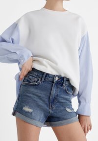 Personne portant un short en denim bleu retroussé avec des patchs usés et un pull blanc avec des manches rayées bleues, main sur la hanche.